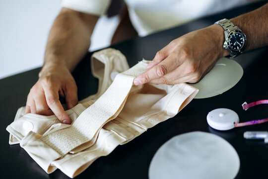 Doctor's hands holding a post-operative compression bra, with silicone breast implants and measuring tape on the table