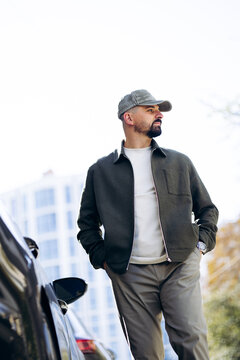 Stylish man in cap and jacket stands by a car on a sunny city street, looking away with a confident, thoughtful expression