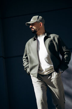 Man in baseball cap and green jacket standing confidently, hands in pockets, against a dark wall in strong sunlight