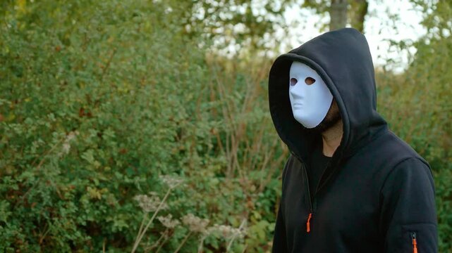 Criminal in white mask walking beside woodland in black hooded clothing. Masked murderer and violent offender concept among trees. Killer, rapist, maniac, crime and potential victim theme in rural