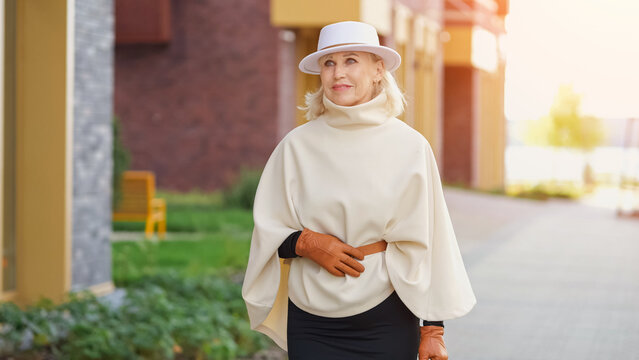 Stylish woman wears white wide brim hat and flowing top while standing on city walkway. Fashion portrait conveys poised confidence, with brown gloves adding polished seasonal touch.