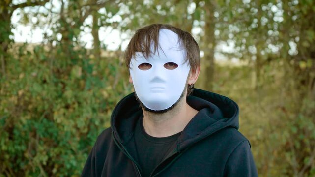 Maniac in white face mask walking through woodland in dark hoodie. Anonymous criminal and murderer concept with concealed identity. Killer, rapist, violent crime, victim and mentally ill offender