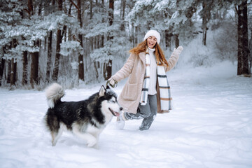 Young woman is having fun in snow-covered forest with Siberian husky. Woman and her dog are out for a walk on winter day, enjoying their time together. Concept of pets, fun, and leisure. © maxbelchenko