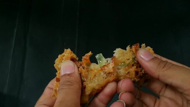 Close up footage of hands breaking apart a crispy Indonesian vegetable fritter (bakwan) against a dark background.