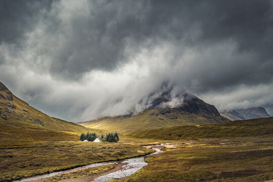 The River Coupall passes by the Wee White House at Glencoe in the Highlands of Scotland with Buachaille Etive Beag shrouded in dark rain clouds behind