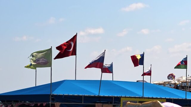 Row of colorful national flags fluttering in steady sea breeze above blue beachfront canopy at sunny seaside resort, flags rising and dipping with wind, bright clear sky, distant umbrellas and water