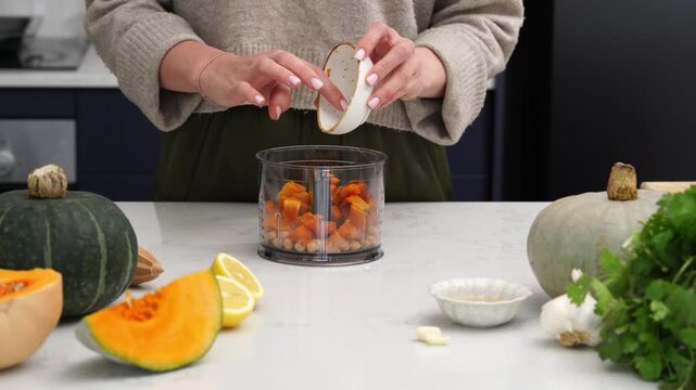 Hands pouring roasted pumpkin and chickpeas into food processor bowl on bright marble countertop in modern kitchen while preparing creamy dressing and mixing ingredients for healthy autumn meal