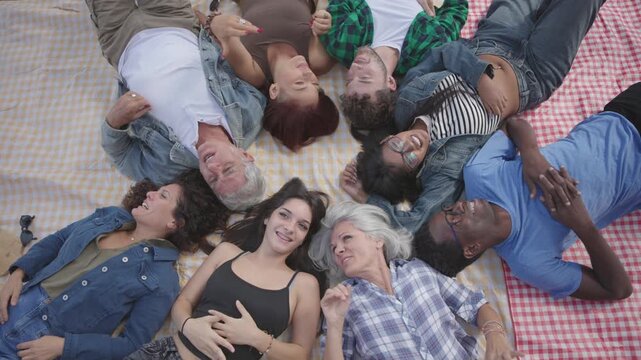 Overhead view of happy multiracial friends lying together on a picnic blanket in a circle, laughing and smiling, showing friendship, unity, diversity, togetherness and summer fun.