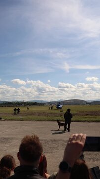 Vertical screen military police dog handler stands with a working dog during a training exercise near a helicopter as spectators watch the demonstration at an airfield event