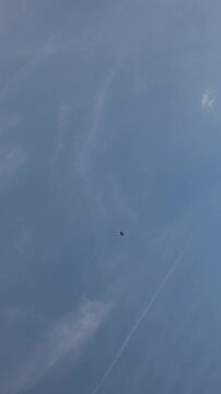 Vertical screen military fighter jet flies high across a vast blue sky leaving a long white vapor trail behind as scattered clouds drift through the clear summer atmosphere