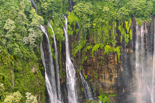 Tumpak Sewu Waterfall in Lumajang, East Java, Indonesia. Hundreds of cascading streams fall from a curved cliff into a lush tropical gorge, surrounded by dense rainforest and rising mist.