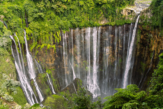 Tumpak Sewu Waterfall in Lumajang, East Java, Indonesia. Hundreds of cascading streams fall from a curved cliff into a lush tropical gorge, surrounded by dense rainforest and rising mist.