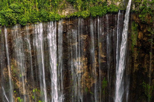 Tumpak Sewu Waterfall in Lumajang, East Java, Indonesia. Hundreds of cascading streams fall from a curved cliff into a lush tropical gorge, surrounded by dense rainforest and rising mist.