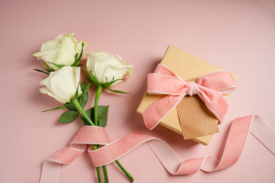 Kraft gift box with white satin ribbon and cream roses on pink background