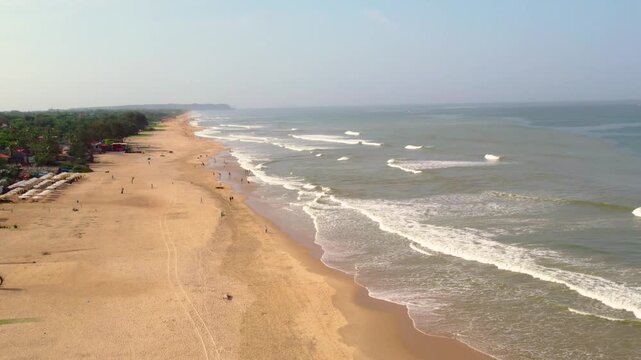 Aerial view of Vagator Beach with gentle waves washing onto the golden sand and two people walking along the shoreline in Anjuna, Goa, India.