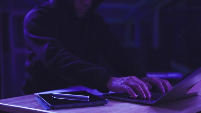 Hooded hacker types on laptop keyboard in dark room beside digital tablet. Cybersecurity threat attack, malware breach infiltration, network security espionage, programming cybercrime surveillance.