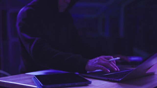 Hooded hacker operates digital tablet beside laptop computer inside dark room. Cyber security breach investigation, network vulnerability penetration testing, digital espionage threat protection.