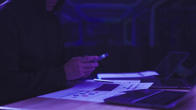 Hooded hacker types on mobile phone beside laptop documents in dark office room. Cybercrime threat monitoring, data breach espionage prevention, network security vulnerability investigation.