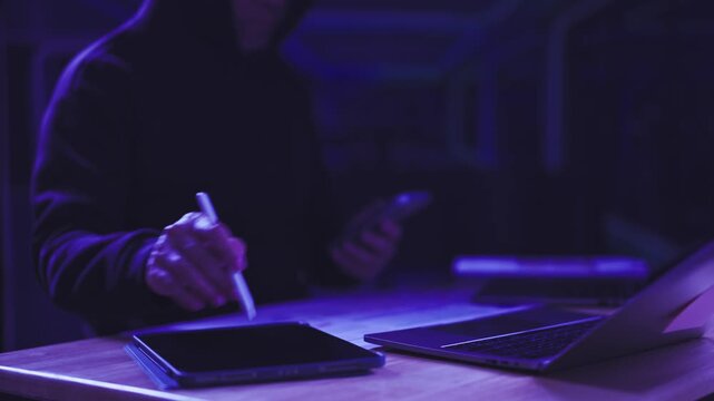 Hooded hacker operates digital tablet beside laptop computer in dark neon room. Cybercrime threat detection, online fraud prevention, unauthorized data breach investigation, information security.