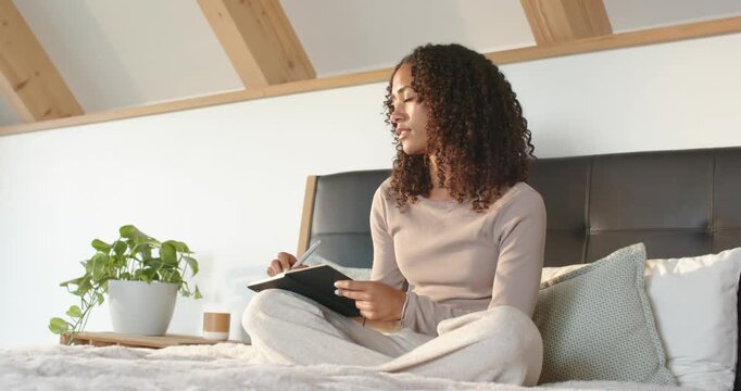 African American woman holding notebook on lap, sitting on bed writing with pen, pausing to plan