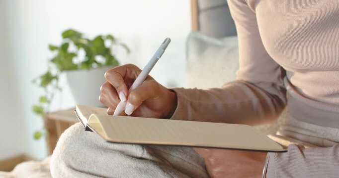 Right hand is writing in lined notebook on couch as pen is touching paper, journaling