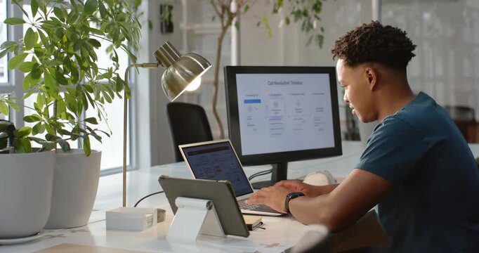 African American man focusing at desk on dual screens, typing, updating slide, glancing up, smiling