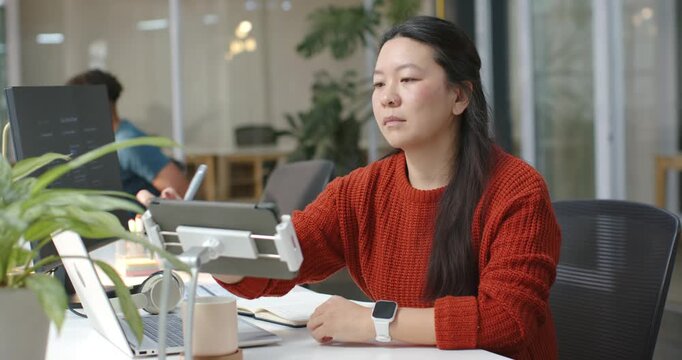 Asian woman in rust-red sweater adjusting tablet on stand, referencing laptop and writing notes
