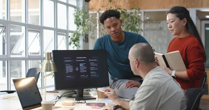 Diverse coworkers gathering and reviewing printouts, monitor at office desk, gesturing and planning