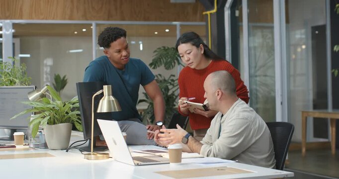 Diverse team leaning, taking notes while seated man pointing at laptop, explaining papers at office