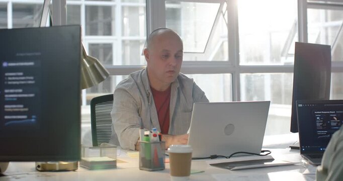 Glancing at right monitor, man sitting in bright office, typing laptop and taking notes for later