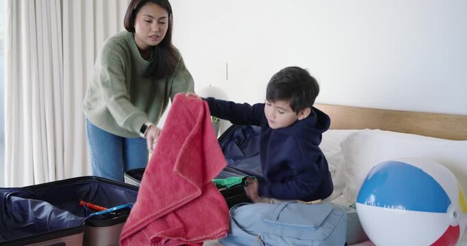 Asian mother and son packing open suitcase on bed, folding red towel while preparing for trip