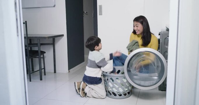Asian mother and son sorting clothes, opening dryer door, passing garments for folding at home