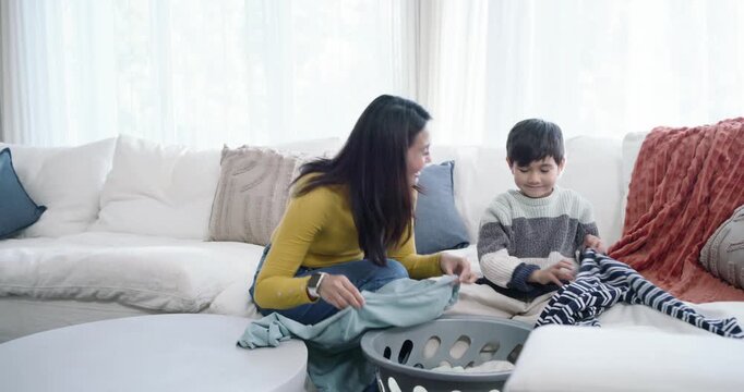 Diverse mom and child folding blue-top, striped-sweater on couch by gray basket, placing on ottoman