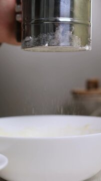 Close up of flour cascading from a metal hand sifter into a white mixing bowl. A woman prepares cinnamon roll dough from scratch in her home kitchen.