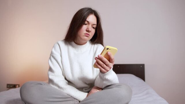Emotionless woman scrolling social media while sitting on bed. Slow motion footage portrays doomscrolling and mental exhaustion from excessive screen time.
