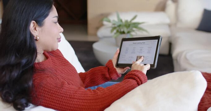 Woman wearing red knit holding tablet, tapping and swiping on sofa in living room confirming lights