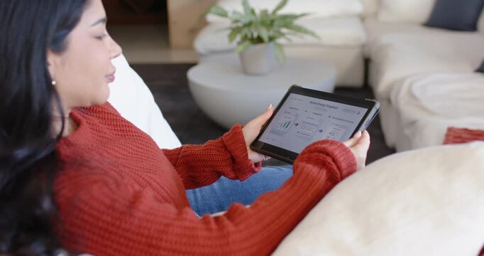 Woman sitting on sofa in rust sweater while checking energy on tablet tapping screen, copy space