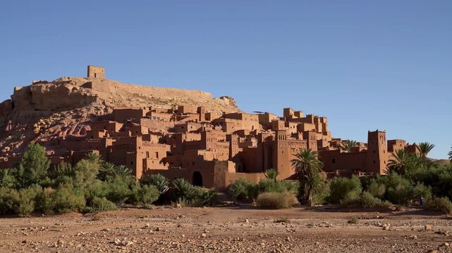 Ait Ben Haddou near Ouarzazate, Morocco, the fortified mud brick ksar rising on a hillside with towers, earthen houses and a granary on top, palm trees and shrubs in front