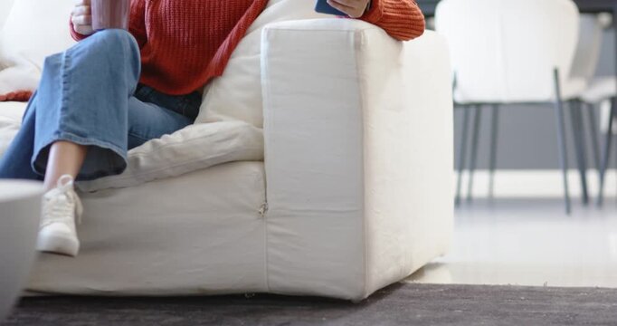 Woman sitting in living room in rust sweater, holding mug, tapping phone, starting robot on rug