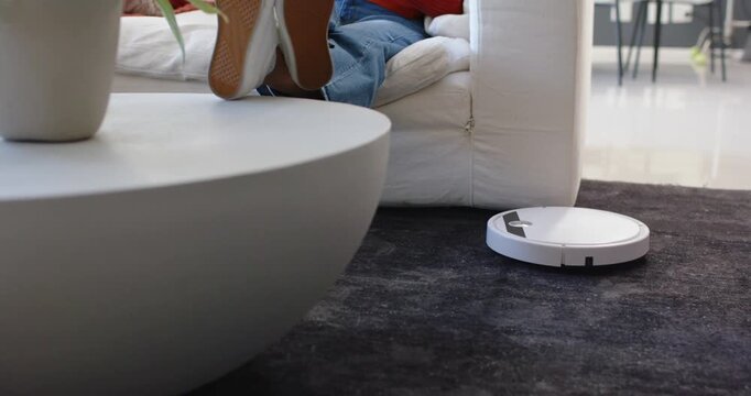Woman reclining on sofa in red sweater, jeans, robot vacuum passing, reaching remote