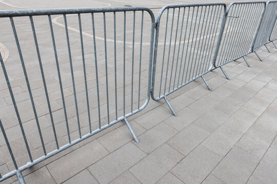 Metal crowd control barriers line a paved urban area, forming a temporary barricade for event safety and pedestrian management. Repeating vertical bars and neutral tones create an industrial scene