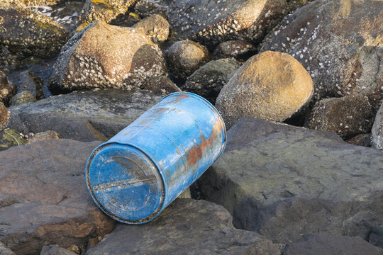 A weathered blue barrel lies on dark coastal rocks near barnacle-covered boulders, suggesting marine debris, shoreline pollution and human impact on the ocean and coastal environment