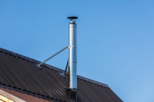 A shiny stainless steel chimney flue rises from a sloped metal roof, secured by support brackets against a clear blue sky, highlighting modern residential ventilation, roofing details