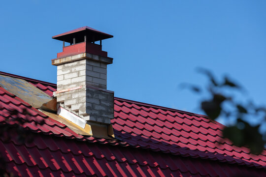 Close-up of a red metal roof and brick chimney framed by soft foreground foliage, highlighting residential architecture, roof textures and lines under a bright clear blue sky on a sunny day