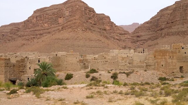 Abandoned mud brick ksar near Ouarzazate, Morocco, with high crumbling clay walls and small windows at the foot of red rock cliffs in a dry gorge, desert shrubs in front