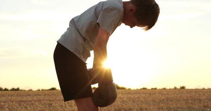 Young boxer rests bent forward in harvested field at sunset. Boy wearing boxing gloves recovers after outdoor workout. Warm sunlight creates calm mood for resilience and fitness stories.
