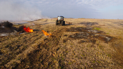 An elevated aerial drone photograph showing a blue agricultural tractor driving across a wide harvested field during controlled burning. © Andrey