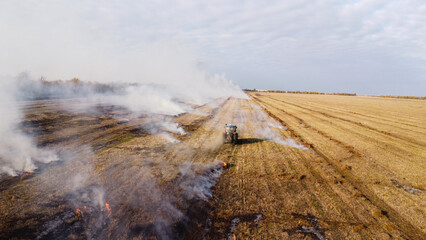 An elevated aerial drone photograph showing a blue agricultural tractor driving across a wide harvested field during controlled burning. © Andrey