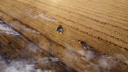 An elevated aerial drone photograph showing a blue agricultural tractor driving across a wide harvested field during controlled burning. © Andrey