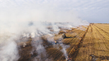 An elevated aerial drone photograph showing a blue agricultural tractor driving across a wide harvested field during controlled burning. © Andrey